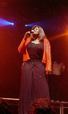 A woman wearing a sparkling black outfit and orange jacket. Her eyes are closed and she is holding a microphone in one of her hands.