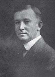 A young man with dark hair wearing a white shirt, dark tie, and dark jacket