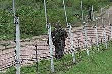 A soldier in camouflage walks next to a gravel road inside a wire fence with white posts.