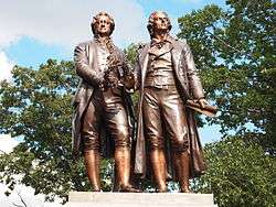 Photograph of the statue of Goethe and Schiller taken from the front. There are trees and blue sky behind them; only the top of a stone pedestal is shown in the photograph.