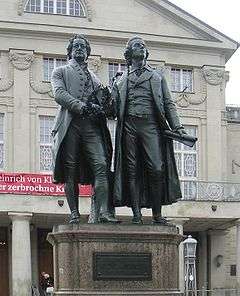 Photograph of a large bronze statue of two men standing side-by-side and facing forward. The statue is on a stone pedestal, which has a plaque that reads "Dem Dichterpaar/Goethe und Schiller/das Vaterland".