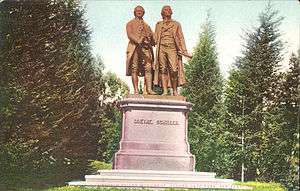 Picture postcard of a large bronze statue of two men on top of a stone pedestal. The pedestal has the engraved words "Goethe. Schiller." on its front face. The statue is surrounded by tall trees. There is printing along the bottom of the postcard that says, "1671 - Goethe Schiller Monument, Golden Gate Park, San Francisco, California" printed along the bottom.