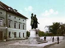 A bronze statue of two men stands on a stone pedestal. The statue is in the middle of a city square; on the left is the facade of a building. Three people are standing in the square and looking at the statue. The bronze statues are noticeably larger than life-sized.