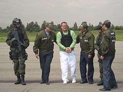 A handcuffed man in a light green T-shirt, flak jacket and white pants being escorted by two men on either side in green jackets and caps with the Spanish word "Policia" identifying them as police officers. To the left is a soldier in body armor and helmet, carrying an assault rifle. At the right is another police officer, his back to the camera, slightly obscuring another similarly equipped soldier.