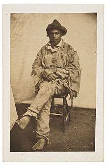 Full length, black-and-white photograph of a seated African-American. He is barefoot and wearing very ragged clothing, having escaped slavery