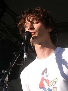 Brown-haired man wearing a white shirt singing to a microphone