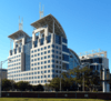 Ground-level view of a 12-story building and 9-story building, connected by an arched glass atrium; the facade is tan with dark blue glass windows. The roofline of the building is curved at several points, and two twin spires extend from the roof on the anterior side of the building.