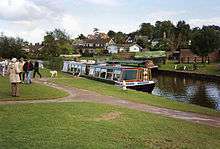A narrow boat moored alongside grassy area. Houses can be seen in the background.