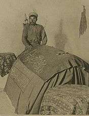 A sepia-toned photograph showing a man wearing a turban, his left arm cradling a thin staff, and standing behind and with his right hand upon a centotaph draped with fabric on which is an inscription in Arabic script