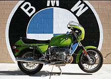 Green BMW R90S motorcycle parked in front of a brick wall which is painted with a large BMW logo