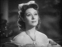 Black-and-white still of a woman gazing upwards, wearing an off-the shoulder dress, a prominent necklace, and a bow in her dark hair.