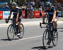 A pair of cyclists on the road, wearing matching black jerseys with steel blue trim. Spectators are visible on the roadside and behind barricades.