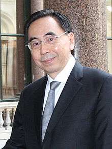 a smiling with wavy hair, wearing glasses, a suit and a blue tie while looking straight at the camera