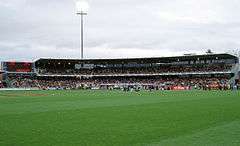 A two tier stand and scoreboard filled with people in the backdrop of an oval grass playing surface.