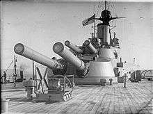 Two large gun turrets seen from the deck of a battleship; each turret has two long guns