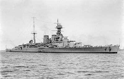 A front-quarter view of a large gray warship with two masts, two funnels, and four visible gun turrets at anchor