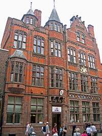 The front of a building with three storeys plus attics is seen from a slight angle.  The ground floor is in stone; the upper storeys are in brick with stone dressings.  On the front is a frieze containing heraldic shields, a coat of arms, and a variety of windows; at the top are two turrets and a shaped gable.