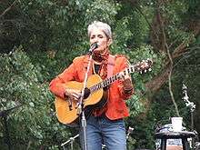 Baez plays in blue jeans and orange waist jacket, against a backdrop of lush trees