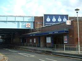 A red-bricked building with a blue sign reading "HAINAULT STATION" in white letters and a lamp post in front all under a blue sky with white clouds