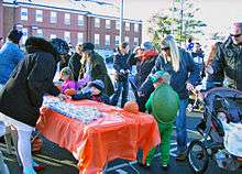Children receiving small treats from a table with a shiny orange covering and two small pumpkins on it. One child to the right is in costume as a green turtle. There are adults behind them wearing heavy coats and hats. In the rear is a large brick building with snow on the roof