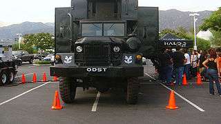 A large, grey truck sits in a parking lot, surrounded by orange cones. People file into the truck via a queue on one side. The truck is branded with "Halo" and "ODST" markers.