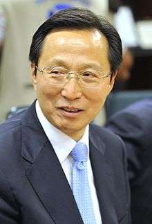a smiling man with a wavy haircut looking leftwards, wearing glasses, a white shirt, a suit and a blue tie with black dots