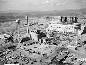 An aerial view of the Hanford B-Reactor site from June 1944. At center is the reactor building. Small trucks dot the landscape and give a sense of scale. Two large water towers loom above the plant.