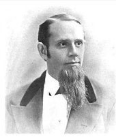 A middle-aged American gentleman of the immediate post-civil-war era. In this black-and-white portrait shot the subject looks to the viewer's right. His hair is short and sharply combed, and a beard is prominent on his chin. He wears a dark suit and white shirt.