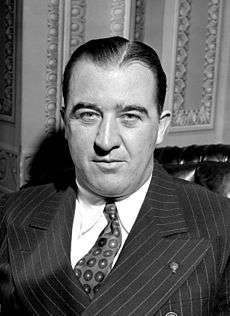 A man with black hair wearing a white shirt, black pinstriped jacket, and patterned tie