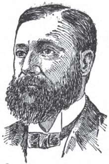 A man with dark hair and a bushy mustache and beard wearing a black jacket and tie and white shirt