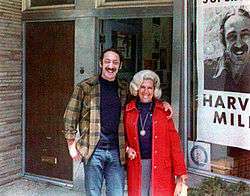 A color photograph of Milk with long hair and handlebar moustache with his arm around his sister-in-law, both smiling and standing in front of a storefront window showing a portion of a campaign poster with Milk's photo and name