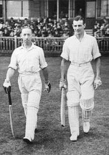 Two small men aged around 30 walk off the steps of a cricket ground pavilion onto the grass towards the centre of the playing area. Both wear light-coloured shirts, with rolled up sleeves, trousers and pads to protect their legs, all white. They wear batting gloves and hold a bat in readiness for play. The crowd sit behind a wooden fence; most are wearing suits and black cylindrical hats.