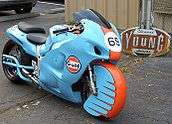 A pale blue motorcycle with a prominent Gulf Oil logo and a unique front fender that encloses almost the entire front wheel on asphalt with a sign in the background that says Young Choppers and Hot Rods.