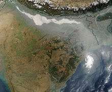 Satellite view of the northern portion of South Asia. The arc of a substantial range of mountains, the Himalayas, dips into the shot, then back up out of view. Immediately beneath, a large region of plains is hidden by a continuous cloud-like opaque mass that has collected along the southern margins of the mountains. It proceeds eastward, staying just south of the Himalayas, then bends due south to reach the Bay of Bengal. Two parts of the mass appear particularly dense, showing up as bright white blobs in the shot.