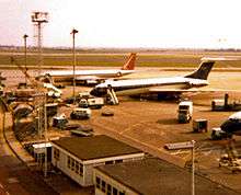 A South African Airways Boeing 707 in former orange, blue and white livery in the background at London Heathrow Airport, parked next to a BOAC Vickers VC10.