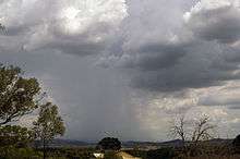 The sky is covered with dark clouds, from the middle of which a shaft of rain or hail comes down on hills on the horizon.