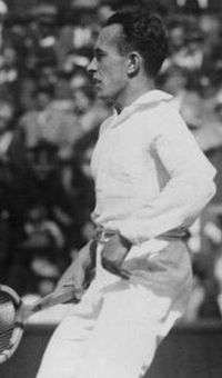 A man in white pants and a white shirt steps back, holding a wooden racket in his right hand