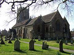 Stone building with arched windows and square tower partially obscured by trees. In the foreground are gravestones.