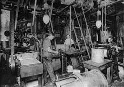 Photograph of a large workshop crowded with machinery. There are at least four men working there. There are belts coming down from the ceiling that drive the machinery. Two electric lighting fixtures are also hung from the ceiling.