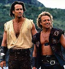 Two men, the one on the left has long brown hair and is taller than the one on the right. The man on the right has long blond hair and is wearing a tunic top and leather trousers and a gauntlet on his right arm. The man on the right is wearing a waistcoat, leather trousers and a medallion around his neck.