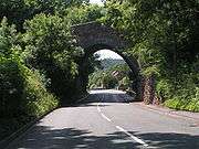 A view along a road, looking through a stone bridge that is partly obscured by trees.