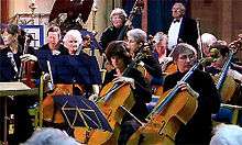 A selected view of an orchestra playing, cellos at the front, double basses behind.