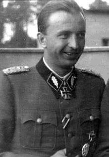 A black-and-white photograph of a smiling man in semi profile wearing a military uniform and a neck order in shape of an Iron Cross.