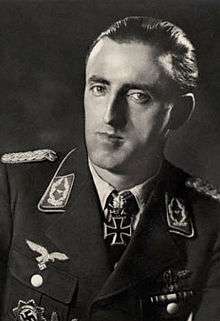 The head and shoulders of a man, shown in semi-profile. He wears a military uniform with various military decorations and an Iron Cross at the front of his shirt collar. His hair is dark and short and combed back, his nose is long and bent and his mouth is thin; he is looking into the camera