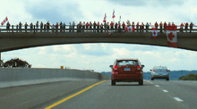 A bridge showcased against the sky, with the ground not visible. Lining the bridge are people, some holding Canadian flags.