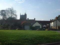 Church tower seen arising behind stone buildings with tile roofs, one of which has a pub sign. Foreground is grass