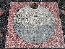 Moon Landing monument, with square pink terrazzo surround (not the usual charcoal color), with light gray terrazzo Moon disk showing TV emblem at top and the brass lettering "Neil A. Armstrong, Edwin E. Aldrin and Michael Collins, 7/20/69, Apollo XI"