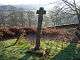 A tail, plain, short-armed cross on a plain base in a graveyard overlooking a trees and a green valley