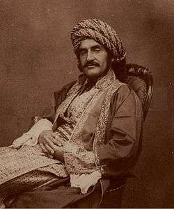 Sepia photograph of a man in 19th century Middle Eastern dress, with a large moustache, reclining in a chair with his hands crossed across his lap