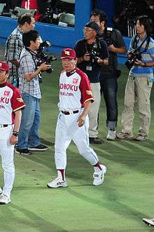 Japanese man wearing a white and red uniform walking onto a baseball field. Several photographers are standing behind him.